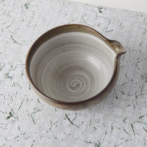 May include: A white ceramic bowl with a brown rim and a swirling gray design. The bowl has a spout on one side.