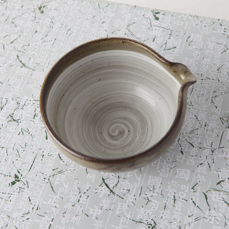 May include: A white ceramic bowl with a brown rim and a swirling gray design. The bowl has a spout on one side.