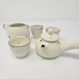 Puede incluir: Un juego de té de cerámica blanca con un acabado moteado. El juego incluye una tetera con asa, una jarra y tres tazas.
