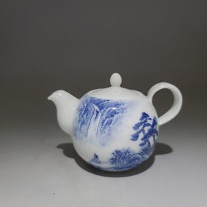 May include: A white porcelain teapot with a blue and white landscape design. The teapot has a round body and a small spout. The lid is also decorated with a blue and white design.