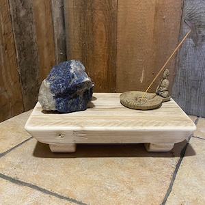 May include: A wooden platform with a blue and white stone, a small ceramic incense burner with a seated figure, and a lit incense stick. The platform has a natural wood finish and rests on four legs.