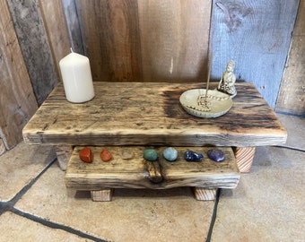 May include: A rustic wooden altar with two tiers, featuring a white candle, a small Buddha statue, a ceramic incense holder, and colorful stones.
