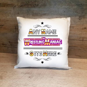 May include: White pillow with the text "ANY NAME THE WRESTLING MANIAC SITS HERE" in black and gold lettering. The word "MANIAC" is in purple and has a gold outline. There is a gold star above the text and a gold star below the text.