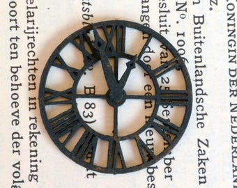 May include: A black metal clock face with Roman numerals and a black metal hour and minute hand. The clock face is 7.6 cm in diameter.