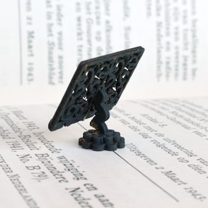 May include: A miniature, decorative black metal stand featuring an openwork square design. It has a curved stem and a circular base. The stand is designed to hold small objects and is placed on a book.