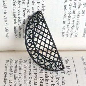 May include: A black metal bookmark with a decorative, intricate design. The bookmark is shaped like a half-circle and features a repeating pattern of squares and swirls.