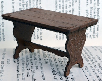 May include: A miniature wooden table with a dark brown finish and intricate carvings on the legs. The table has a rectangular top and is sitting on a book.