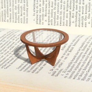 Puede incluir: Una mesa de madera en miniatura con una tapa de vidrio. La mesa es marrón y tiene un diseño único y moderno de mediados de siglo. La mesa está sobre un libro.