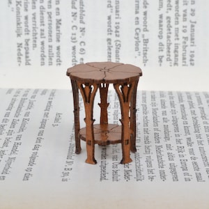 Op de afbeelding: Een miniatuur houten tafel met een bruine afwerking en ingewikkelde houtsnijwerk. De tafel heeft een rond blad en twee planken.