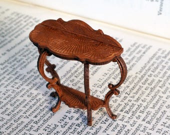 May include: A miniature wooden table with a brown finish and intricate carvings. The table has a rectangular top and two curved legs. The table is sitting on a book.
