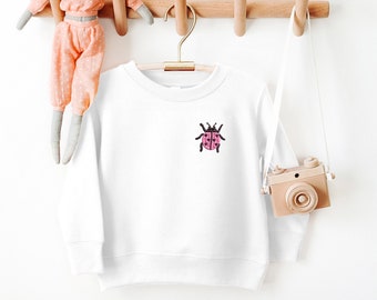 May include: White sweatshirt with a pink ladybug embroidered on the front. The sweatshirt is hanging on a wooden rack with a stuffed animal elephant nearby.