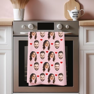 May include: A pink kitchen towel with a repeating pattern of faces and red hearts. The faces are of a man and a woman. The towel is hanging on an oven door. A white teapot with the text "LOVE U LOTS GIFTS" is on the counter.