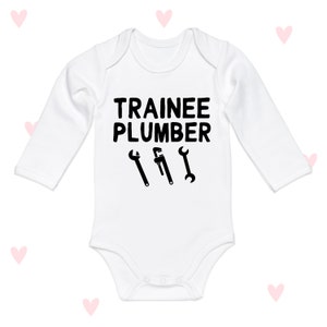 Puede incluir: Mono blanco de manga larga para bebé con texto negro que dice "Trainee Plumber" y tres ilustraciones de llaves inglesas negras.