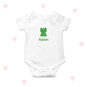Puede incluir: Un body blanco para bebé con una rana verde y el nombre "Ruben" bordado en la parte delantera.
