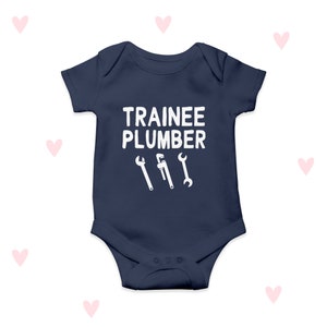 Puede incluir: Un body de bebé azul marino con texto blanco que dice "Trainee Plumber" y tres ilustraciones de llaves blancas.