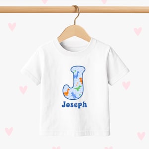 Op de afbeelding: Wit T-shirt met een blauwe letter "J" gevuld met een dinosauruspatroon in oranje, groen en blauw. De naam "Joseph" is in blauw onder de letter gedrukt. Het shirt hangt aan een houten hanger.