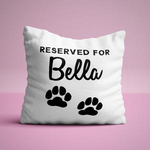 May include: White pillow with black text that reads "Reserved for Bella" and two black paw prints.