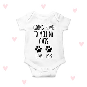 May include: White baby onesie with black text that reads "Going home to meet my cats" with two black paw prints below the text. The paw prints have the names "Luna" and "Pops" written below them.