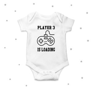 Op de afbeelding: Witte baby romper met zwarte tekst die "Player 3 is loading" zegt en een zwart-witte illustratie van een videogamecontroller.