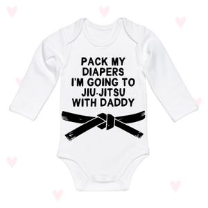 Peut inclure: Body bébé blanc à manches longues avec le texte noir "PACK MY DIAPERS I'M GOING TO JIU-JITSU WITH DADDY". Un motif de ceinture noire est en dessous. Le body a un col rond et des boutons-pression.