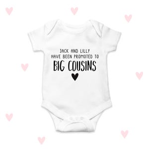 Può includere: Body da bambino bianco con la scritta nera "JACK AND LILLY HAVE BEEN PROMOTED TO BIG COUSINS", con un cuore nero sotto. Il body ha maniche corte e bottoni automatici sul fondo. Cuori rosa sparsi sullo sfondo.