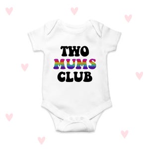 May include: White baby bodysuit featuring the text "TWO MUMS CLUB" in black and rainbow hues. The bodysuit has short sleeves and a snap closure. Pink heart shapes are scattered around the bodysuit.