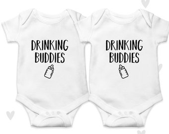 May include: Two white baby onesies with short sleeves. The onesies have black text that says "Drinking Buddies" and a black outline of a baby bottle.