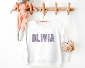 May include: White sweatshirt with a purple outline and a colorful floral pattern spelling out the name "OLIVIA".