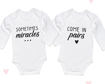 May include: Two white long-sleeved baby onesies with black text. One onesie says "Sometimes miracles..." and the other says "Come in pairs" with a small black heart.