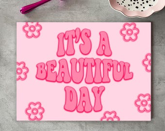 May include: A pink rectangular sign with the text "IT'S A BEAUTIFUL DAY" in bubble letters. The sign has a light pink background with pink flower graphics. A pink spoon and a decorative plate are in the background.