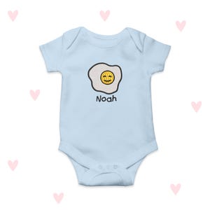 May include: A pale blue baby bodysuit featuring an embroidered smiling fried egg. The egg has a yellow yolk and a white outline. The name "Noah" is printed below the egg in black. The bodysuit has short sleeves and poppers at the bottom.