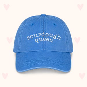 May include: A bright blue baseball cap featuring the text "sourdough queen" embroidered in white thread across the front panels. The cap has a curved brim, a button on the top, and stitched eyelets for ventilation on each of the six panels.
