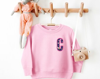 May include: Pink sweatshirt with a colorful letter C patch on the front. The letter is surrounded by a pink outline and has a white background with a pattern of stars and unicorns.