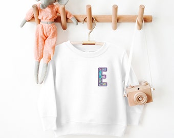 May include: A white sweatshirt with a colorful, patterned letter "E" on the front. The sweatshirt is hanging on a wooden rack with a toy elephant and a toy camera. The design features shades of blue, purple, and pink.