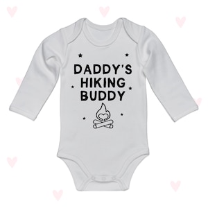 Op de afbeelding: Witte babybody met lange mouwen met de zwarte tekst "DADDY'S HIKING BUDDY". Het ontwerp bevat een kampvuurafbeelding en kleine steraccenten. De body is gemaakt van zacht materiaal en heeft drukknoopsluitingen.