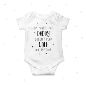 Puede incluir: Body de bebé blanco con el texto "I'M PROOF THAT DADDY DOESN'T PLAY GOLF ALL THE TIME" en negro. El body tiene mangas cortas y cierre de broches.
