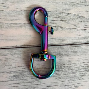 May include: A multi-coloured metal swivel clasp with a spring-loaded gate. The clasp has a rainbow iridescent finish, with hues of purple, blue, green, and gold. The clasp is designed to attach to a leash or other hardware.