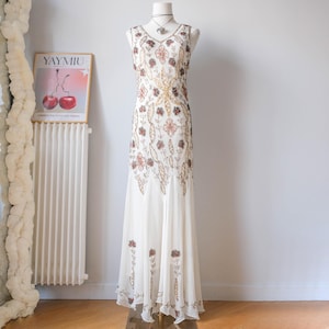 Peut inclure: Une robe longue blanche sans manches ornée de perles complexes dans des tons or, bronze et cuivre. La robe présente un motif floral et végétal sur tout le corsage et la jupe, avec une encolure en V et un ourlet évasé et fluide.