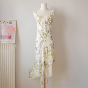 Può includere: Un abito midi senza maniche di colore bianco con un motivo floreale nei toni del giallo e del grigio. L'abito presenta uno scollo a V con bordo arricciato sul corpetto e un orlo a balze a cascata che crea una silhouette asimmetrica.