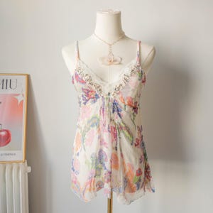 May include: A sleeveless camisole top featuring a cream base with a vibrant multicoloured floral and paisley print. The garment has a V-neckline trimmed with white lace and clear decorative beads, thin spaghetti straps, and a flowing hemline.