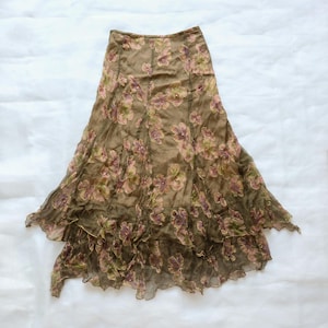 May include: A midi-length skirt in a muted olive green tone featuring a semi-sheer overlay with a floral pattern in shades of pink, purple, and green. The skirt has a high-waisted fit, vertical seam detailing, and a layered, ruffled hemline.