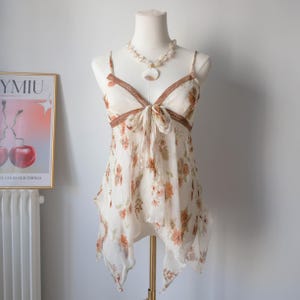 Peut inclure: Un caraco crème orné d'un motif floral orange et marron, présenté sur un mannequin. Il est doté de fines bretelles, d'une bordure en dentelle marron et d'un nœud à la poitrine. Un collier de coquillages complète l'ensemble.