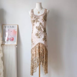 May include: A vintage-style, blush-colored dress with intricate gold floral embroidery and a delicate lace hem. The sleeveless dress is displayed on a mannequin, showcasing its elegant design. A gold necklace adds a touch of glamour to the ensemble.