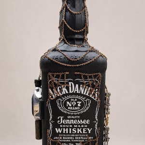 Peut inclure: Une bouteille de whisky Jack Daniel's noir mat ornée de chaînes couleur cuivre drapées autour du goulot et d'un motif de chaîne en toile sur l'étiquette avant. Une montre-bracelet argentée est fixée sur le côté avec une petite chaîne.