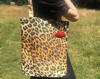 May include: A tote bag with a leopard print pattern in shades of brown and black. The bag has a gold-colored strap and a red lips keychain attached. The background is a grassy field.