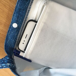 以下が含まれることがあります： 白いジッパー付きポケットが付いた青いデニムのトートバッグ。白いスマートフォンと青いペンがポケットに入っています。