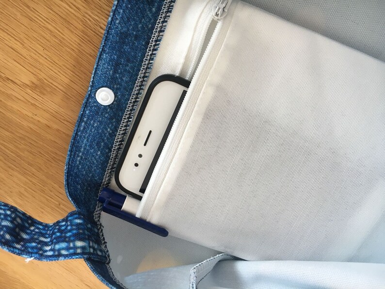 以下が含まれることがあります： 白いジッパー付きポケットが付いた青いデニムのトートバッグ。白いスマートフォンと青いペンがポケットに入っています。
