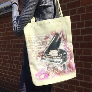 以下が含まれることがあります： クリーム色のトートバッグ。水彩画風のグランドピアノのイメージが描かれています。ピアノは黒とグレーで描かれ、楽譜とピンクの花のアクセントがあります。バッグはシンプルなデザインで、ショルダーストラップが付いています。