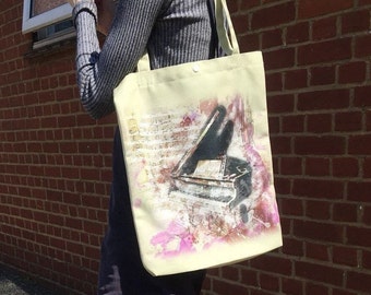 May include: Cream-colored tote bag featuring a watercolor-style image of a grand piano. The piano is depicted in shades of black and gray, with musical notes and pink floral accents. The bag has a simple design with a shoulder strap.