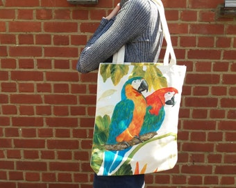 May include: A white tote bag featuring a colorful illustration of two parrots perched on a branch. The parrots have blue, orange, and red plumage. The bag has cream-colored handles and a botanical background.